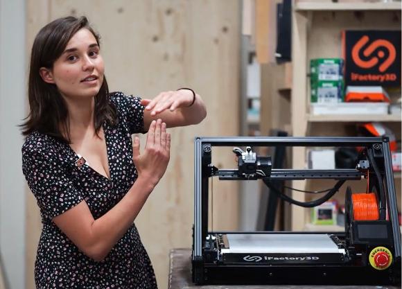 A young woman stands next to the ONE PRO assembly line printer in a workshop and explains, gesticulating a bit