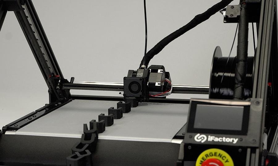 Der Fließband Drucker One Pro von iFactory3D bei der seriellen additiven Fertigung vieler kleiner schwarzer Bauteile, einige haben sich schon über den Schaber vom Belt gelöst.