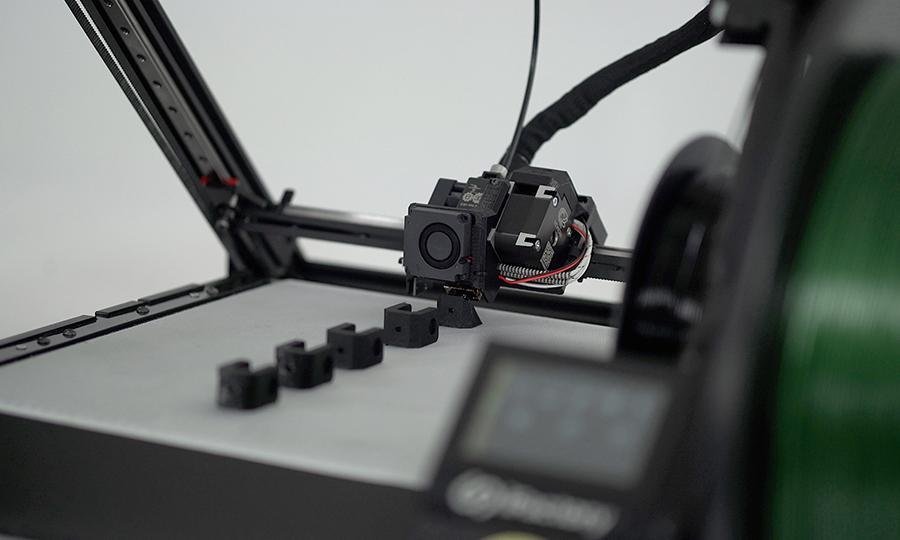 Der iFactory3D One Pro beim Druck einer Kleinteilserie aus schwarzem PETG Material, seitlich in den Bauraum fotografiert. Rechts verschwommen sind Filamentrolle und Display zu erkennen, im Fokus das silberne Druckbett mit Druckkopf.