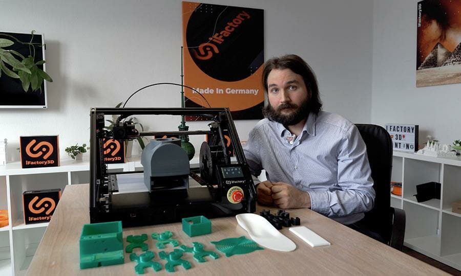 Martin Huber sitzt im Büro neben  einem One Pro Drucker. Davor liegt eine Vielzahl verschiedener Objekte, die auf dem 3D Fließband Drucker hergestellt wurden.
