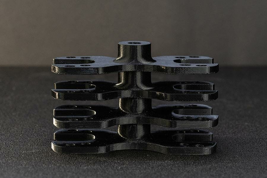 Hier sind vier gleiche Bauteile aus schwarzem PETG gestapelt, mittig erhaben mit je vier Schraublöchern an den Seiten und einer großen zentralen Ausbuchtung.