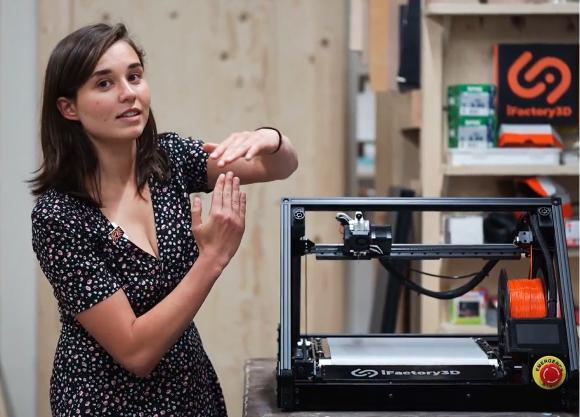 Eine junge Frau steht neben dem ONE PRO Fließband Drucker in einer Werkstatt und erklärt etwas gestikulierend