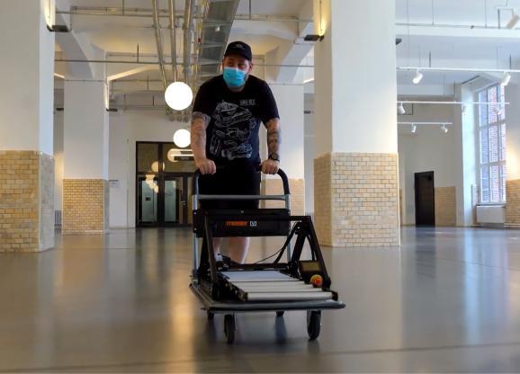 Ein Mann mit Tattoos und Tshirt schiebt den ONE PRO samt montierter Rollenverlängerung auf einem Rollwagen durch einen großflächigen, leeren Flur mit industriellem Charme