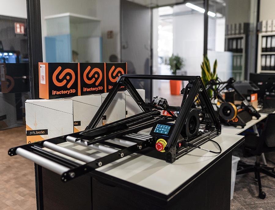 The One Pro in an office setting, with filament boxes from 3dk.Berlin and iFactory3D set up next to it. On the One Pro including extension, a long component for a musical instrument can be seen in print, made of black PETG.