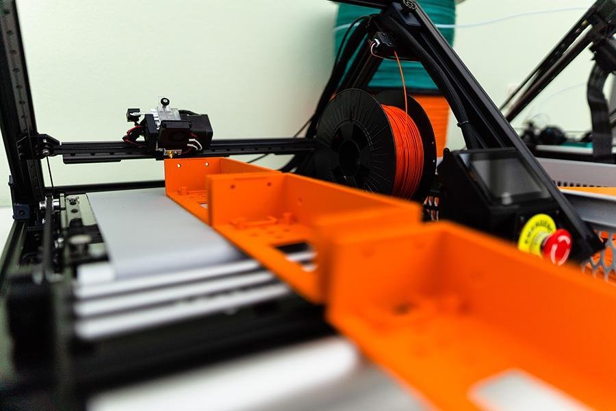 Der 3D Fließband Drucker One Pro direkt neben einem weiteren Drucker, mit 3 großen, in orangem PETG gedruckten Displayverkleidungen, die über die Rollenverlängerung aus dem rechten Bildrand laufen