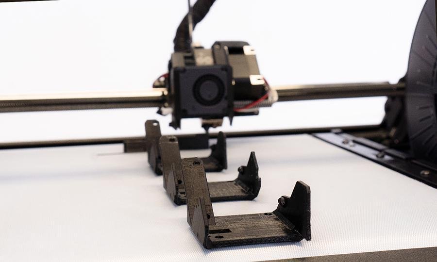 Close-up of 3 black print parts on the conveyor belt print bed of the One Pro, focus on front print part, in the background print head creating another identical object
