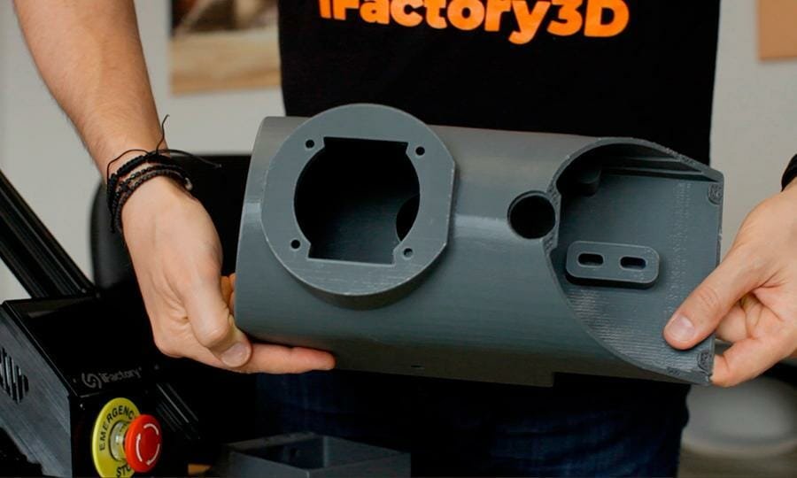 3D gedruckte Vorrichtung eines Schaltgetriebes wird hochgehalten von Person mit iFactory3D T-Shirt Aufschrift, daneben Notausknopf des One Pro zu erkennen. Das Schaltgetriebe ist hohl mit einigen Aussparungen und Löchern, und aus grauem Kunststoff gefertigt