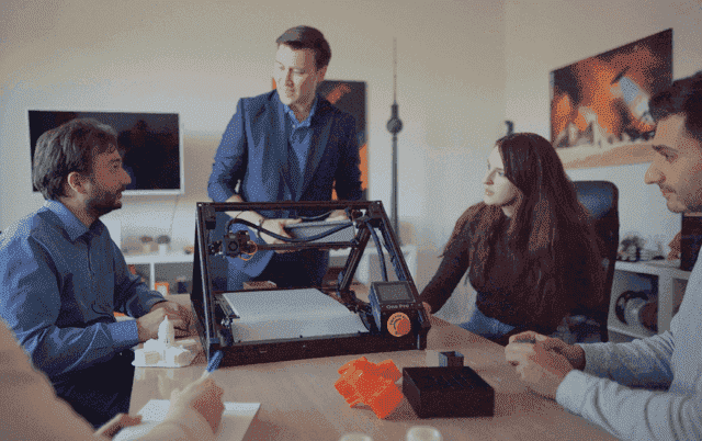 iFactory3D Büro: 5 Personen sind um einen Tisch versammelt, auf dem sich der 3D Fließband Drucker One Pro sowie einige unterschiedliche Druckteile befinden.