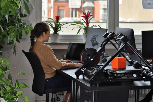 A woman sits in an office working on her computer with a 3D belt printer in the foreground, placed on her desk.