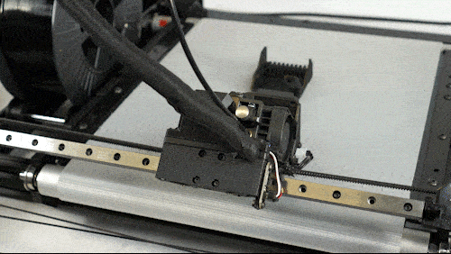 Rear view of the One Pro 3D printer: the  print head is moving from side to side while the guid rail moves up and down on the conveyor belt print bed