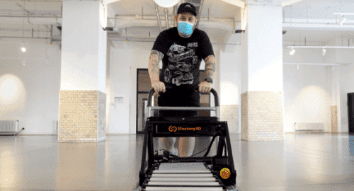 A man with tattoos and a T-shirt pushes the ONE PRO, complete with mounted roller extension, on a trolley through a large, empty hallway with industrial flair