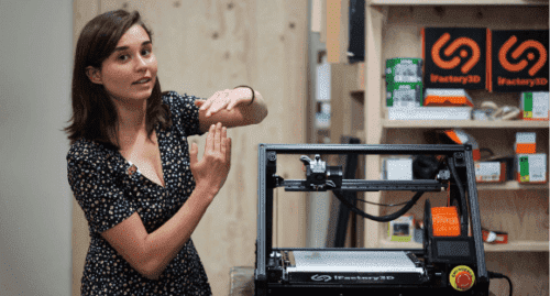 A young woman stands next to the ONE PRO assembly line printer in a workshop and explains, gesticulating a bit