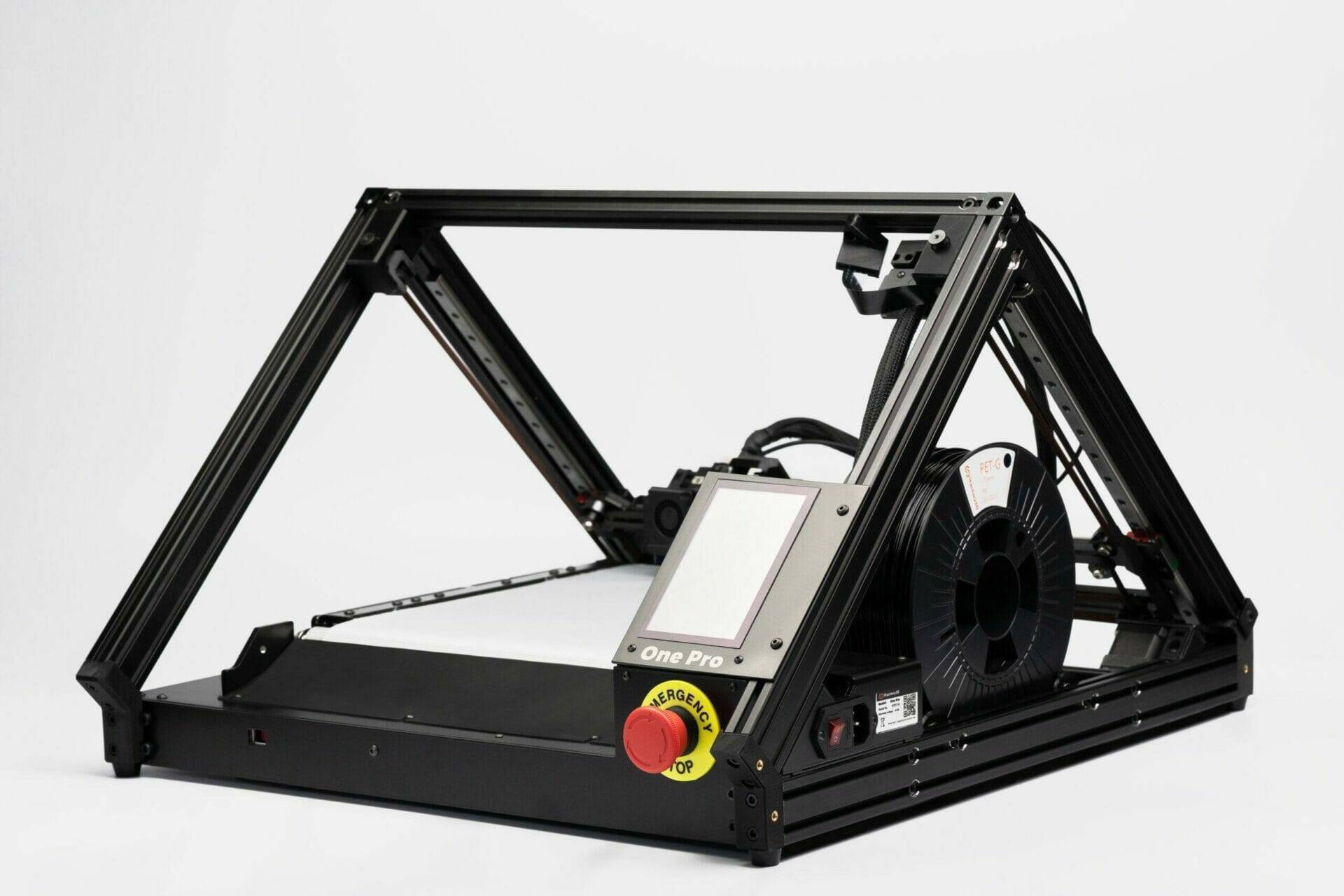 ONE PRO conveyor belt 3D printer with pyramid-like structure, black frame and silver print bed; black filament spool in the build space, which is located directly behind the screen and the emergency stop button