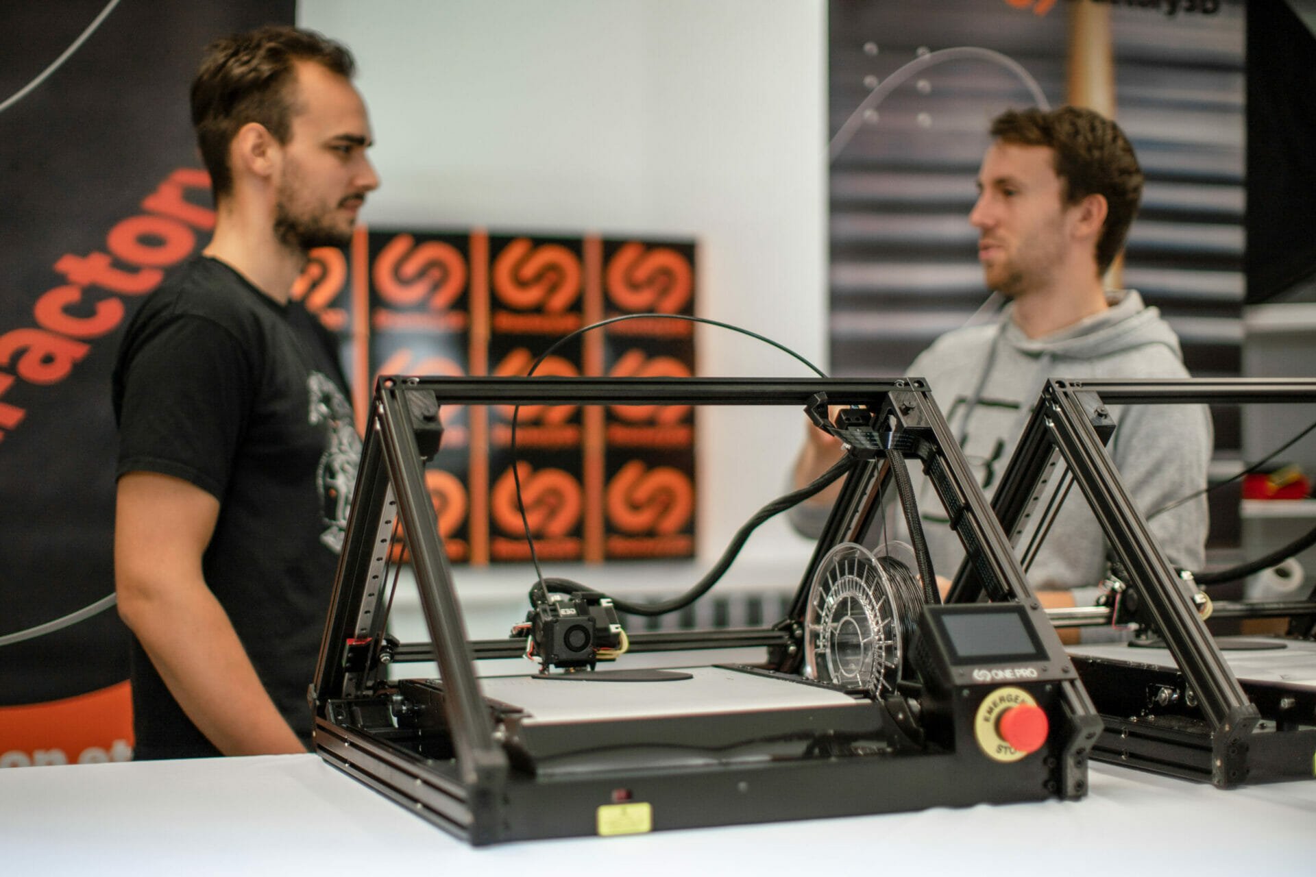 Zwei junge Männer hinter einem Tisch, auf dem mehrere 3D-Fließbanddrucker der Marke iFactory3D zu sehen sind. Das Firmenlogo des Herstellers ist im Hintergrund mehrfach zu erkennen.