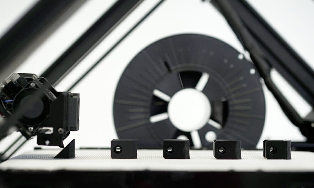 Der One Pro 3D-Fließbanddrucker von der Seite, mit einigen kleinen schwarzen Bauteilen auf der Fließbanddruckfläche