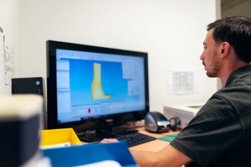 Jemand arbeitet am PC, auf dem Bildschirm ist ein CAD-Programm zu erkennen, in dem eine 3D-Datei einer Beinorthese geöffnet ist.