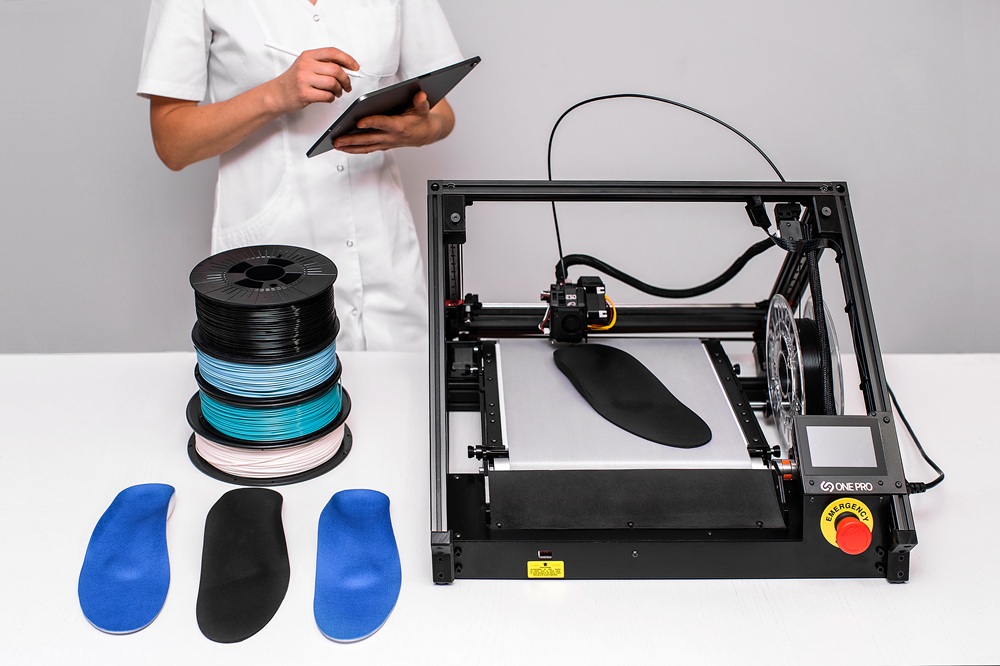 3D-Drucker mit Förderband druckt orthopädische Einlagen, daneben Filamentrollen und eine Person mit Tablet.